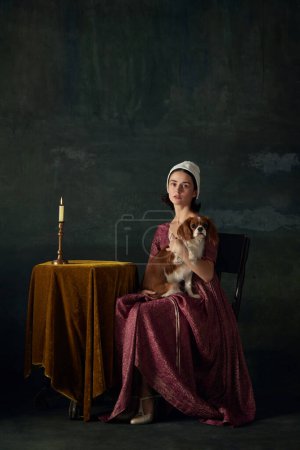 Téléchargez les photos : Jolie et élégante jeune femme à l'image de la demoiselle de la Renaissance assise à table avec un chien mignon sur fond vintage sombre. Concept d'histoire, comparaison des époques, beauté, art, créativité - en image libre de droit