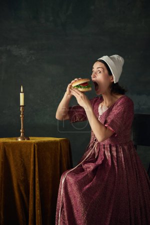 Photo for Junk food lover. Medieval woman in historical costume in image of renaissance maid eating burger against dark green background. Concept of history, comparison of eras, beauty, art, creativity - Royalty Free Image