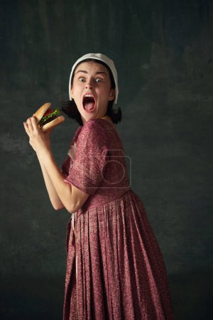 Foto de Mujer medieval en traje histórico, vestido con la imagen de la criada renacentista comer hamburguesa emocional sobre fondo verde oscuro. Concepto de historia, comparación de épocas, belleza, arte, creatividad - Imagen libre de derechos