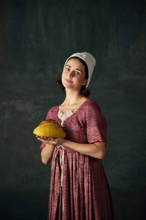 Téléchargez les photos : Boulangerie. Portrait de jolie jeune femme à l'image d'une servante de la Renaissance tenant du pain fraîchement cuit sur fond vert vintage. Concept d'histoire, comparaison des époques, beauté, art, créativité - en image libre de droit