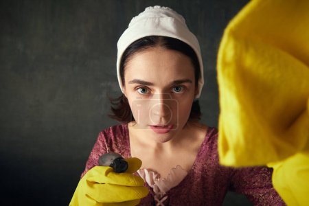 Photo for Close-up portrait of young woman in image of renaissance maid, wearing rubber gloves and cleaning wipes on dark vintage background. Concept of history, comparison of eras, beauty, art, creativity - Royalty Free Image
