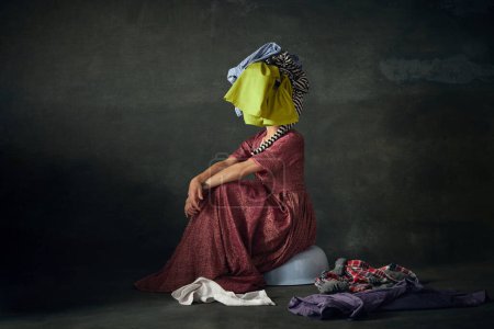 Foto de Mujer en traje de criada medieval sentada en el suelo con la ropa en la cabeza contra el fondo verde vintage. Servicio de limpieza. Concepto de historia, comparación de épocas, belleza, arte, creatividad - Imagen libre de derechos