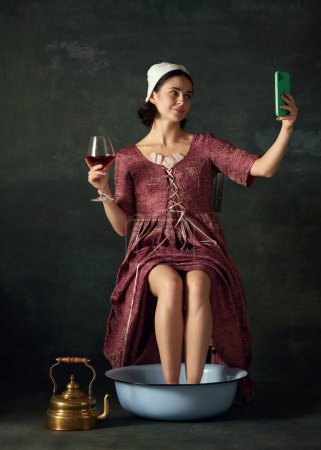 Photo for Woman in renaissance dress, maid sitting with legs into bowl, drinking wine, takign selfie on phone against vintage green background. Concept of history, comparison of eras, beauty, art, creativity - Royalty Free Image