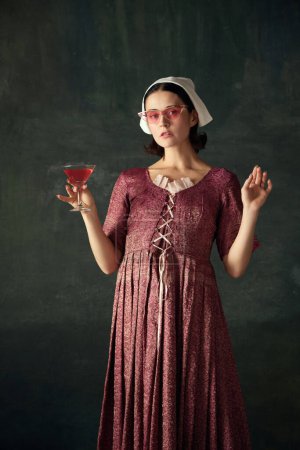 Photo for Portrait of pretty young woman in image of medieval prison, maid sandign in glasses with cocktail against dark vintage background. Concept of history, comparison of eras, beauty, art, creativity - Royalty Free Image