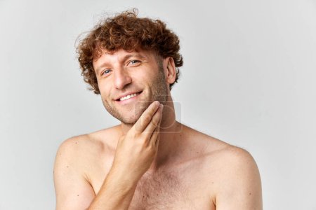 Photo for Time for shaving. Mature smiling man with curly hair holding hand on cheeks against grey studio background. Concept of mens beauty, body and skin care, cosmetology, wellness, ad - Royalty Free Image