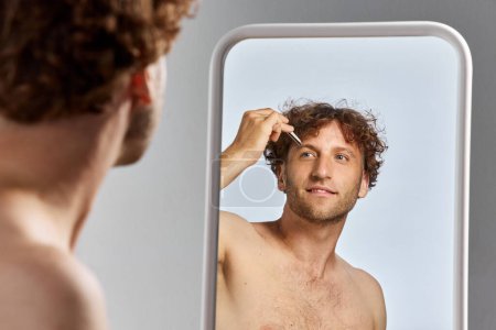 Foto de Hombre maduro guapo mirando en el espejo y tirando de pinzas cejas con fondo gris estudio. Concepto de belleza para hombre, cuidado del cuerpo y la piel, cosmetología, bienestar, ad - Imagen libre de derechos