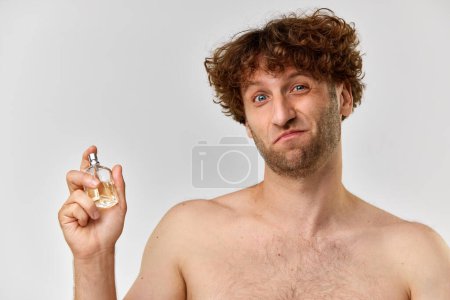 Téléchargez les photos : Portrait d'homme mature torse nu aux cheveux bouclés appliquant du parfum sur fond de studio gris. Concept de beauté masculine, soins du corps et de la peau, cosmétologie, bien-être, publicité - en image libre de droit