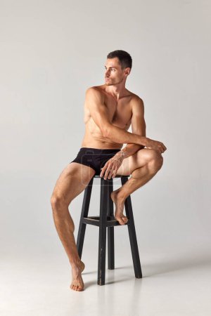 Portrait of muscular, athletic young man sitting on chair shirtless in underwear and posing against grey studio background. Concept of mens health and beauty, body care, fitness, wellness, ad