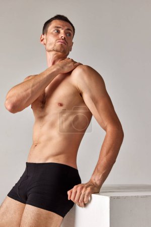 Téléchargez les photos : Portrait d'un bel homme au corps musclé et athlétique posant torse nu en sous-vêtements sur fond de studio gris. Concept de santé et de beauté masculine, soins du corps, fitness, bien-être, publicité - en image libre de droit