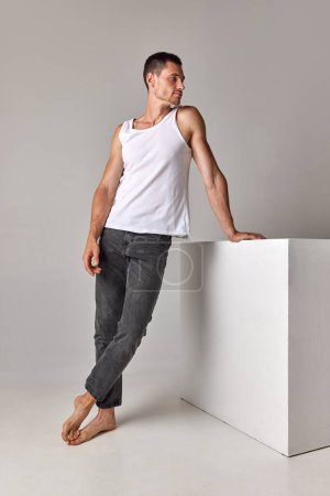 Full-length image of young guy, handsome man posing in singlet and jeans against grey studio background. Concept of mens health and beauty, body care, fitness, wellness, ad