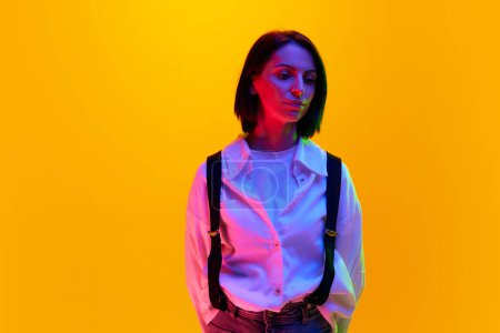 Photo for Portrait of woman in her 30s wearing white shirt, posing with calm emotionless face against orange studio background in neon light. Concept of human emotions, fashion, lifestyle, facial expression, ad - Royalty Free Image