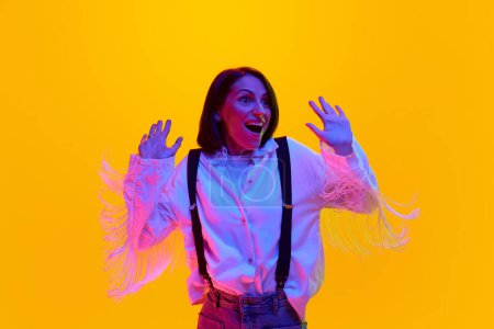 Photo for Happy and excited woman in white shirt and suspenders, standing with shocked face against orange studio background in neon light. Concept of human emotions, fashion, lifestyle, facial expression, ad - Royalty Free Image