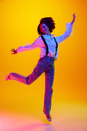 Photo for Full-length image of young woman in her 30s, wearing casual clothes and jumping against orange studio background in neon light. Concept of human emotions, fashion, lifestyle, facial expression, ad - Royalty Free Image