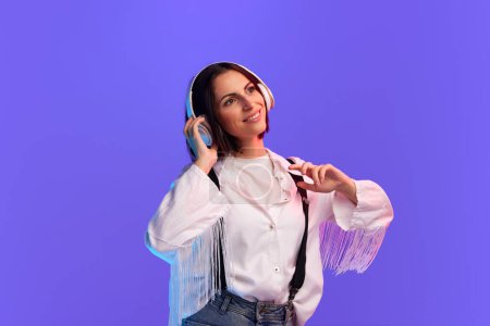 Photo for Beautiful, smiling woman in stylish clothes listening to music in headphones against purple studio background in neon light. Concept of human emotions, fashion, lifestyle, facial expression, ad - Royalty Free Image