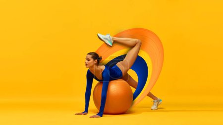 Photo for Stretching. Beautiful young girl in blue bodysuit training with fitness ball onyellow background with abstract design element. Contemporary art collage. Concept of sport, action and motion, health - Royalty Free Image