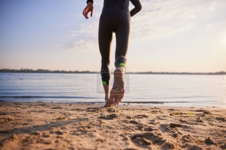 Photo for Sportive man in wetsuit running on beach into water for early morning training. Swimming workout. Cropped image. Concept of professional sport, triathlon preparation, competition, athleticism - Royalty Free Image