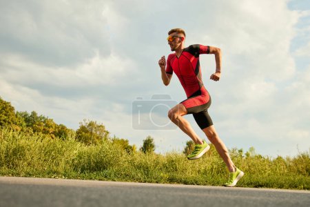 Téléchargez les photos : Athlète professionnel, homme en tenue de sport s'entraînant à l'extérieur par une chaude journée ensoleillée. Courir le long de la route. Concept de sport professionnel, préparation au triathlon, compétition, athlétisme - en image libre de droit