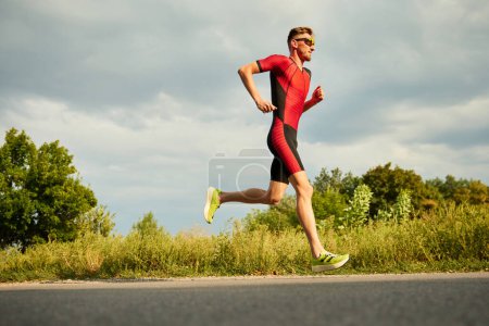 Téléchargez les photos : Athlète professionnel, homme en tenue de sport s'entraînant à l'extérieur par une chaude journée ensoleillée. Courir le long de la route. Concept de sport professionnel, préparation au triathlon, compétition, athlétisme - en image libre de droit