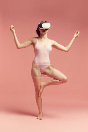 Téléchargez les photos : Yoga virtuel. Femme en body, portant des lunettes vr et des exercices de yoga d'entraînement sur fond de studio rose. Concept de soins du corps et de la peau, fitness, beauté naturelle, santé, technologie moderne - en image libre de droit