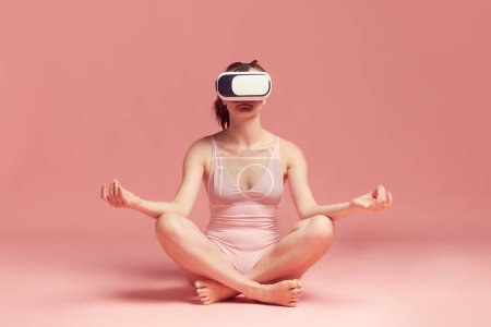 Téléchargez les photos : Méditation en ligne et yoga. Femme en body, portant des lunettes vr et assise sur fond de studio rose. Concept de soins du corps et de la peau, fitness, beauté naturelle, santé, technologie moderne - en image libre de droit