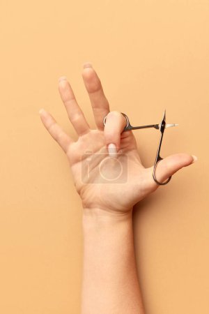 Photo for Female hand with nude, natural looking nails, manicure holding manicure tool - scissors against beige background. Concept of hand care, cosmetics and cosmetology, spa, natural beauty. Poster, ad - Royalty Free Image
