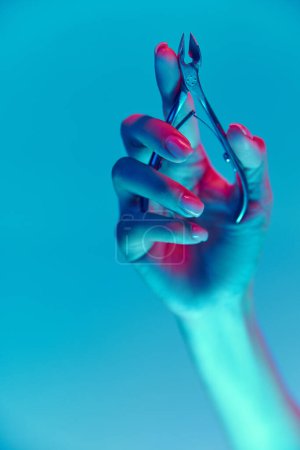 Photo for Female hand with nude, natural nails, manicure holding nippers isolated over blue background in neon light. Concept of hand care, cosmetics and cosmetology, spa, natural beauty. Poster, ad - Royalty Free Image
