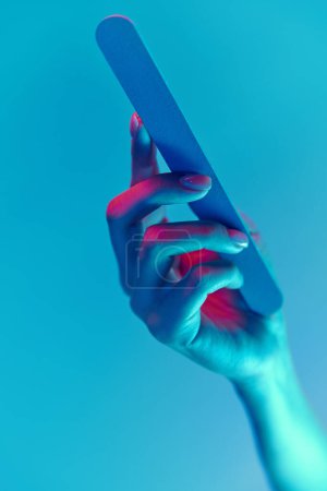 Téléchargez les photos : Main féminine avec ongles nus, naturels, manucure tenant lime à ongles isolée sur fond bleu dans la lumière au néon. Concept de soins des mains, cosmétiques et cosmétologie, spa, beauté naturelle. Affiche, annonce - en image libre de droit