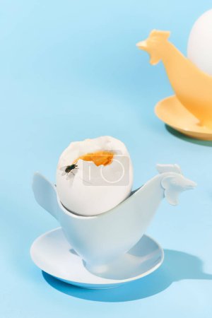Foto de Huevos cocidos en taza de huevo sobre fondo azul claro. Vuela sentado sobre un delicioso huevo. Concepto de desayuno, comida, sabor, salud, creatividad. Fotografía de arte pop. Cartel. Copiar espacio para anuncio - Imagen libre de derechos