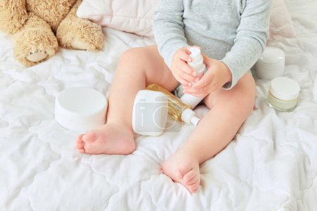 Photo for Cropped image of child legs, toddler sitting in bed with body moisturizing creams, lotions. Skincare products. Concept of childhood, kids cosmetology and natural cosmetics, body care - Royalty Free Image