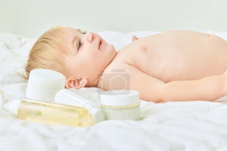 Photo for Happy, lovely child, little baby girl lying on bed at home with skincare products. Moisturizing body with cream. Concept of childhood, kids cosmetology and natural cosmetics, body care - Royalty Free Image