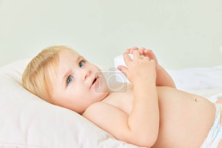 Photo for Cute toddler, little baby girl, child with blue eyes lying in bed and holding moisturizing body cream. Home care. Concept of childhood, kids cosmetology and natural cosmetics, body care - Royalty Free Image