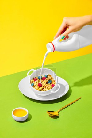 Photo for Morning vibe. Female hand pouring milk into bowl with sweet muesli, cereal with berries against green yellow background. Concept of healthy food, nutrition, pop art, taste. Poster. Copy space for ad - Royalty Free Image