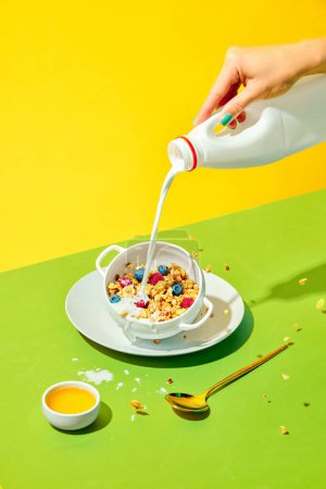 Foto de Desayuno saludable. Mujer vertiendo leche en un tazón con deliciosos muesli, cereal con bayas sobre fondo amarillo verde. Concepto de comida saludable, nutrición, arte pop, gusto. Cartel. Copiar espacio para anuncio - Imagen libre de derechos
