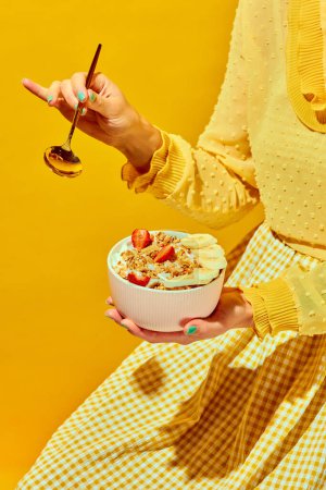 Foto de Mujer en traje agradable sosteniendo cuchara dorada y tazón con muesli, cereal con bayas sobre fondo amarillo. Concepto de comida saludable, nutrición, estilo pop art, sabor. Cartel. Copiar espacio para anuncio - Imagen libre de derechos