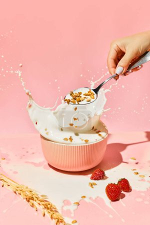 Foto de Cuenco con leche salpicada y mano femenina poniendo muesli, cereal sobre fondo rosa. Desayuno saludable. Concepto de comida saludable, nutrición, estilo pop art, sabor. Cartel. Copiar espacio para anuncio - Imagen libre de derechos