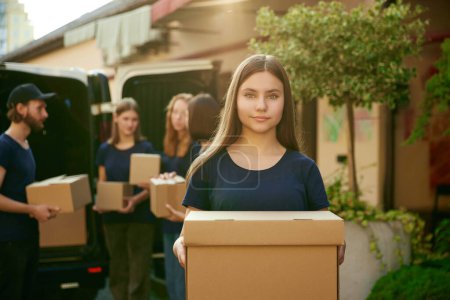 Photo for Focus on girl, volunteer standing outdoors near van on sunny day. Young people on background loading boxes into van. Concept of humanitarian aid, assistance and support, care, social programs - Royalty Free Image