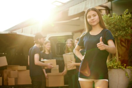 Photo for Focus on girl, volunteer standing near van on sunny day. Young people on background loading boxes into van. Concept of humanitarian aid, assistance and support, care, social programs - Royalty Free Image