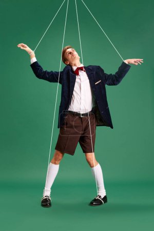 Photo for Social manipulation and pressure. Young guy, marionette on string against green studio background. Concept of human emotions, lifestyle, psychology, social pressure. Ad - Royalty Free Image