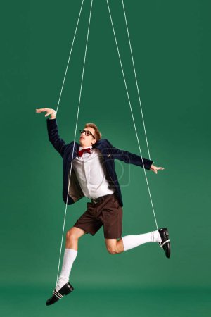 Photo for Young guy, marionette on string in motion against green studio background. Puppet. Pressure and manipulation. Concept of human emotions, lifestyle, psychology, social pressure. Ad - Royalty Free Image