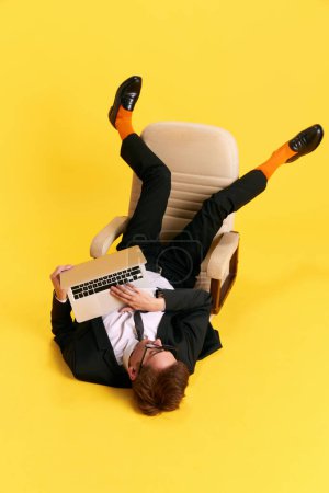 Foto de Una pose inusual. Empresario, freelancer en silla trabajando en portátil contra fondo de estudio amarillo. Empleo. Concepto de emociones humanas, estilo de vida, negocios, ocupación, educación. Anuncio - Imagen libre de derechos