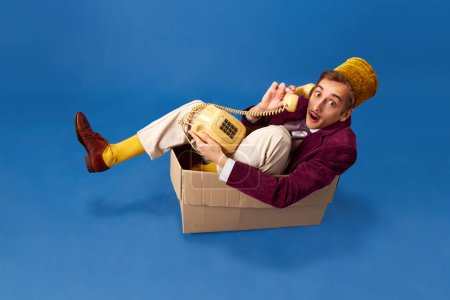 Photo for Young guy, porter sitting in box and talking on retro mobile phone against blue studio background. Concept of human emotions, lifestyle, occupation, employment. Ad - Royalty Free Image