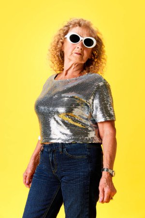 Téléchargez les photos : Élégante femme âgée en haut argenté élégant, jeans et lunettes de soleil posant sur fond jaune studio. Concept d'émotions humaines, mode, personnes âgées, mode de vie, créativité. Publicité - en image libre de droit
