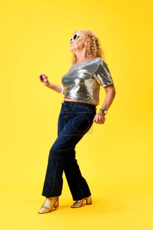 Téléchargez les photos : Longueur totale de femme âgée élégante en haut et jeans étincelants argentés, dansant sur fond de studio jaune. Concept d'émotions humaines, mode, personnes âgées, mode de vie, créativité. Publicité - en image libre de droit