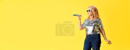 Foto de Senior hermosa mujer en ropa moderna con estilo hablando en el teléfono móvil sobre fondo de estudio amarillo. Concepto de emociones humanas, moda, personas mayores, estilo de vida, creatividad. Anuncio. Banner - Imagen libre de derechos