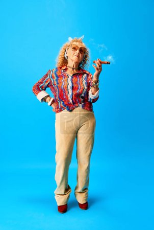 Téléchargez les photos : Hipster. Image pleine longueur de la femme âgée élégante fumant cigare sur fond bleu studio. Concept d'émotions humaines, mode, personnes âgées, mode de vie, créativité. Publicité - en image libre de droit
