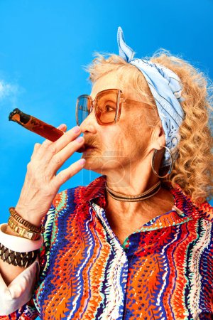 Foto de Retrato de mujer mayor en accesorios elegantes y gafas de sol fumar cigarro sobre fondo estudio azul. Concepto de emociones humanas, moda, personas mayores, estilo de vida, creatividad. Anuncio - Imagen libre de derechos