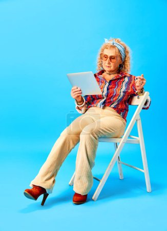 Foto de Senior mujer elegante en ropa elegante sentado en las escaleras y la lectura de noticias en la tableta sobre fondo de estudio azul. Concepto de emociones humanas, moda, personas mayores, estilo de vida, creatividad. Anuncio - Imagen libre de derechos