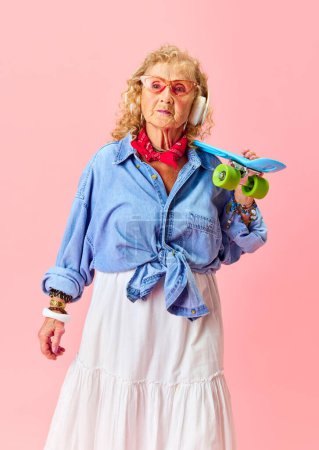 Foto de Un ambiente hipster. Senior hermosa mujer en ropa casual con estilo, camisa, falda y gafas de sol posando con monopatín sobre fondo rosa. Concepto de emociones, moda, personas mayores, estilo de vida. Anuncio - Imagen libre de derechos