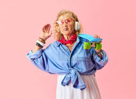 Téléchargez les photos : Ambiance hippie. Senior belle femme dans des vêtements décontractés élégants, chemise, jupe et lunettes de soleil posant avec skateboard sur fond rose. Concept d'émotions, mode, personnes âgées, mode de vie. Publicité - en image libre de droit