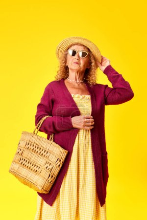 Téléchargez les photos : Elégante dame âgée en beaux vêtements confortables, chapeau de paille et lunettes de soleil posant sur fond jaune studio. Concept d'émotions humaines, mode, personnes âgées, mode de vie, créativité. Publicité - en image libre de droit